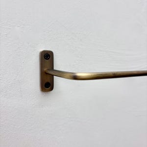 May include: A brass-toned towel bar mounted on a white wall. The bar has a rectangular mounting plate with two screws and a curved, horizontal bar extending outward. The finish has a slightly aged appearance.
