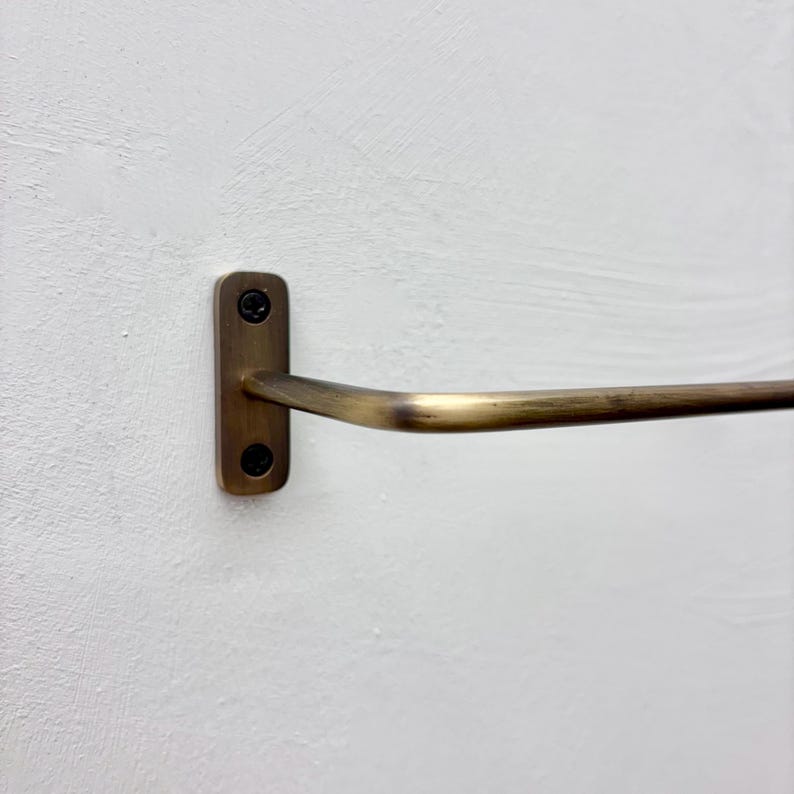 May include: A brass-toned towel bar mounted on a white wall. The bar has a rectangular mounting plate with two screws and a curved, horizontal bar extending outward. The finish has a slightly aged appearance.