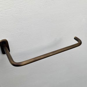 May include: A bronze-colored towel bar mounted on a white wall. The bar has a rectangular base and a curved, horizontal rod. The metal has a slightly aged appearance, suggesting a vintage or industrial design aesthetic.
