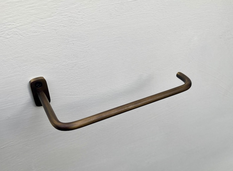 May include: A bronze-colored towel bar mounted on a white wall. The bar has a rectangular base and a curved, horizontal rod. The metal has a slightly aged appearance, suggesting a vintage or industrial design aesthetic.