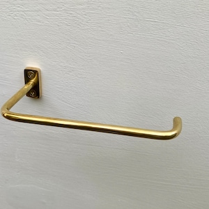 May include: A gold-colored, wall-mounted towel bar. The bar is a simple, modern design with a rectangular mounting plate and a curved, horizontal bar. The bar is made of metal and has a shiny finish. The towel bar is mounted on a white wall.