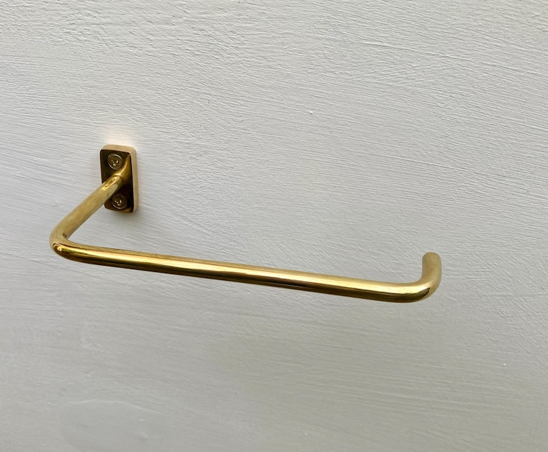 May include: A gold-colored, wall-mounted towel bar. The bar is a simple, modern design with a rectangular mounting plate and a curved, horizontal bar. The bar is made of metal and has a shiny finish. The towel bar is mounted on a white wall.