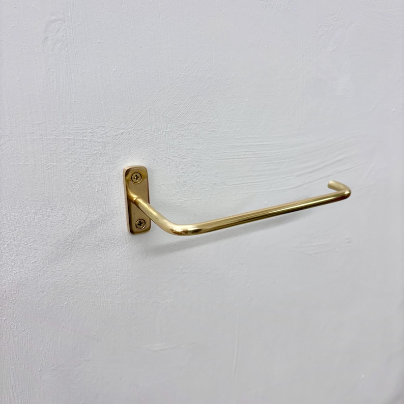 May include: A gold-colored, wall-mounted toilet paper holder. The holder has a rectangular base with rounded corners, secured by four screws. The curved arm extends outward, designed to hold a roll of toilet paper. The fixture is mounted on a white wall.