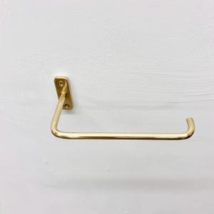 May include: A gold-colored, wall-mounted paper towel holder. The holder has a rectangular mounting plate with two screws and a curved bar for holding the paper towels. The design is minimalist and modern.