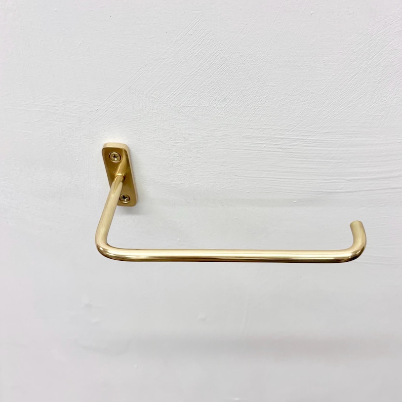 May include: A gold-colored, wall-mounted paper towel holder. The holder has a rectangular mounting plate with two screws and a curved bar for holding the paper towels. The design is minimalist and modern.