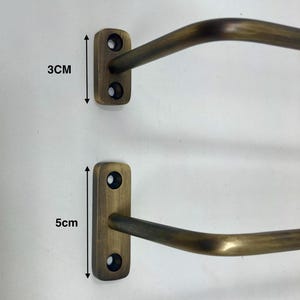 May include: Two antique brass-colored wall-mounted towel racks. The smaller rack is 3 cm (1.2 inches) tall, and the larger rack is 5 cm (2 inches) tall. Each rack has a rectangular mounting plate with two screw holes and a curved bar.