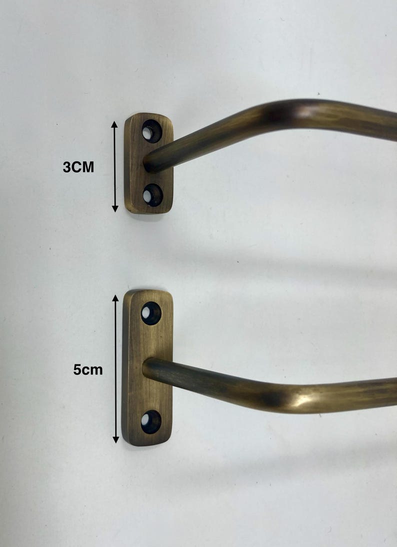 May include: Two antique brass-colored wall-mounted towel racks. The smaller rack is 3 cm (1.2 inches) tall, and the larger rack is 5 cm (2 inches) tall. Each rack has a rectangular mounting plate with two screw holes and a curved bar.