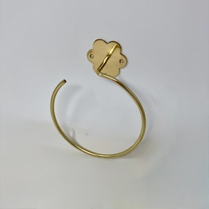 Puede incluir: Un toallero dorado con un soporte de pared en forma de flor. El anillo tiene un diseño simple y curvo, y el soporte de pared tiene forma de flor con pequeños agujeros. El artículo es de metal y está montado sobre una superficie blanca.