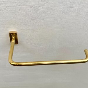 May include: A gold-colored, wall-mounted towel bar. The bar is rectangular at the mounting point and curves into a horizontal bar. The metal has a shiny finish. The bar is designed to hold a towel.