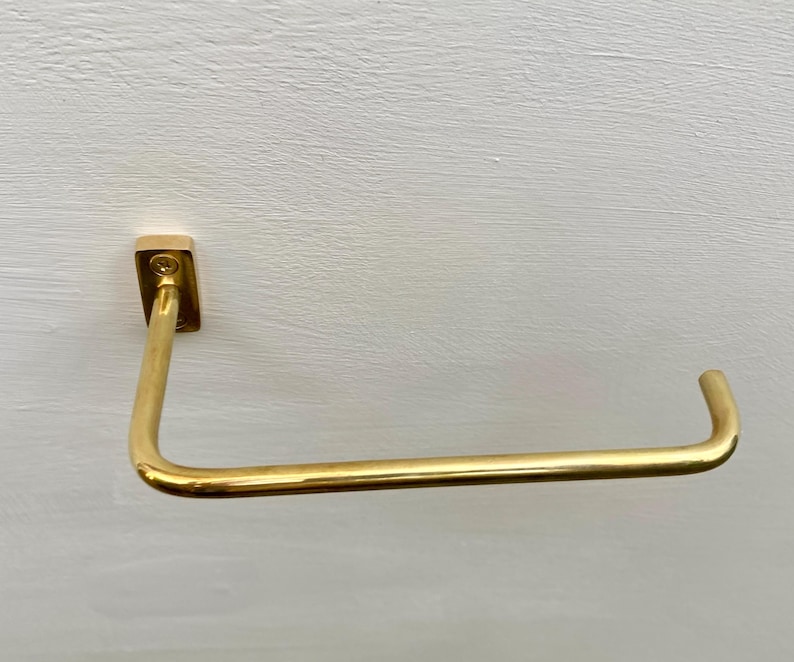 May include: A gold-colored, wall-mounted towel bar. The bar is rectangular at the mounting point and curves into a horizontal bar. The metal has a shiny finish. The bar is designed to hold a towel.