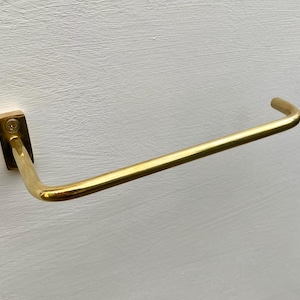 May include: A gold-colored towel bar mounted on a white wall. The bar has a rectangular base and a curved, horizontal arm. The metal has a shiny finish, reflecting light. The design is simple and modern, suitable for a bathroom or kitchen.