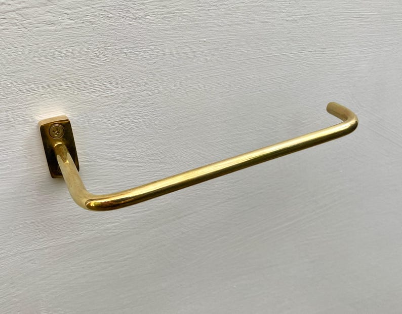 May include: A gold-colored towel bar mounted on a white wall. The bar has a rectangular base and a curved, horizontal arm. The metal has a shiny finish, reflecting light. The design is simple and modern, suitable for a bathroom or kitchen.