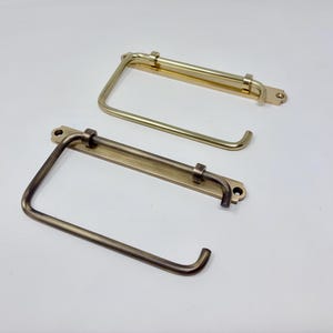 Könnte beinhalten: Zwei Toilettenpapierhalter aus Metall. Einer ist goldfarben, der andere dunkler bronzefarben. Jeder Halter hat eine rechteckige Basis mit zwei Schraubenlöchern und einen gebogenen Arm zur Aufnahme der Rolle. Die Halter sind aus Metall.