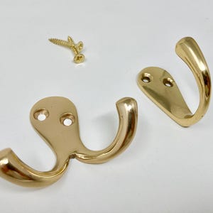以下が含まれることがあります： 白い表面に、真鍮色のコートフック2つと金色のネジ3つ。片方のフックは二重のカーブ、もう片方はシングルフックです。フックにはそれぞれ2つのネジ穴があります。
