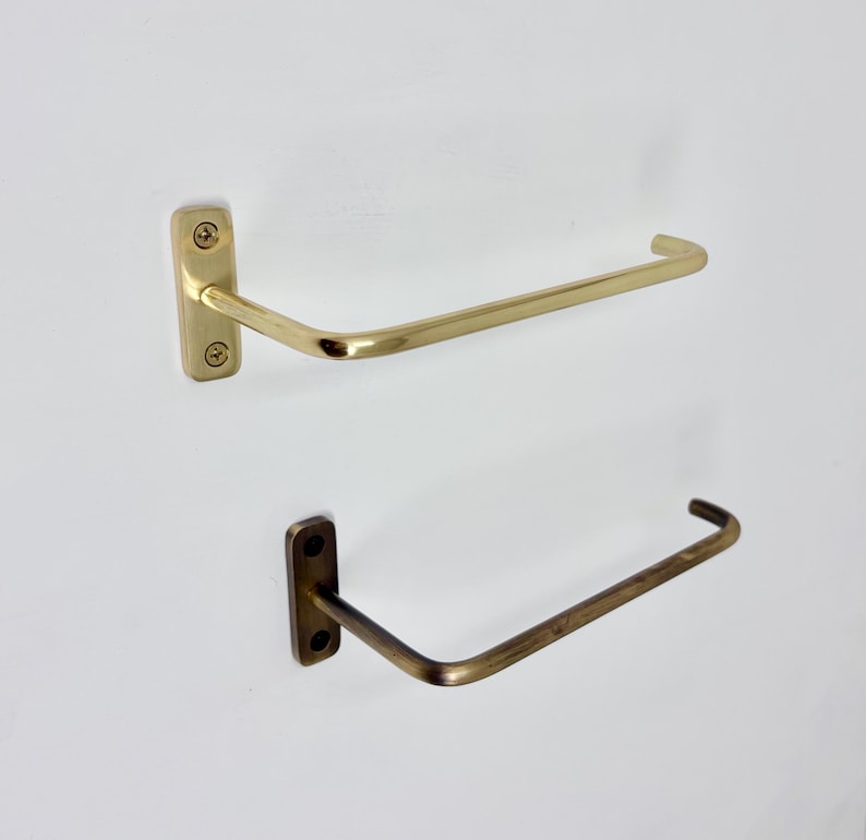 May include: Two metal towel racks. The top rack is a bright gold color, and the bottom rack is a darker, bronze color. Each rack has a rectangular mounting plate with screws and a curved bar for hanging towels. The racks are mounted on a white wall.