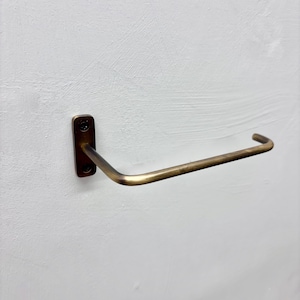 May include: A brass-toned towel bar mounted on a white wall. The bar has a rectangular mounting plate with two screw holes and a curved, horizontal arm. The metal has a slightly aged appearance, suggesting a vintage or antique style.