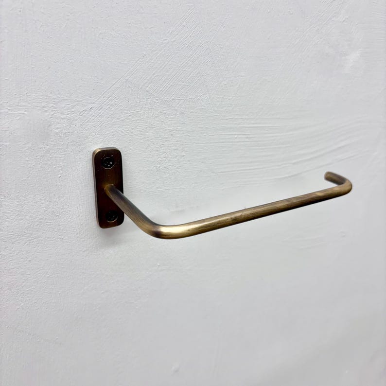 May include: A brass-toned towel bar mounted on a white wall. The bar has a rectangular mounting plate with two screw holes and a curved, horizontal arm. The metal has a slightly aged appearance, suggesting a vintage or antique style.
