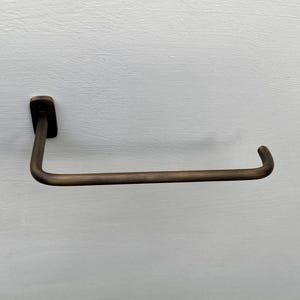 May include: A bronze-colored, wall-mounted towel bar. The bar has a rectangular mounting plate and a curved, horizontal arm. The design is simple and modern, suitable for a bathroom or kitchen.