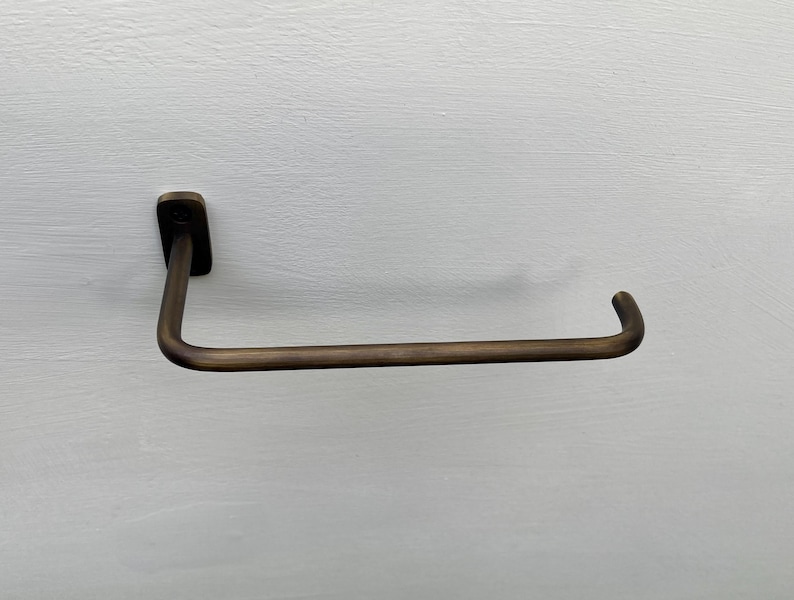 May include: A bronze-colored, wall-mounted towel bar. The bar has a rectangular mounting plate and a curved, horizontal arm. The design is simple and modern, suitable for a bathroom or kitchen.