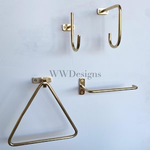 May include: A set of four brass-coloured metal wall fixtures including two J-shaped hooks, one triangular towel ring, and one horizontal towel bar, each mounted with small screws. The text WWDesigns is centred in the middle of the arrangement.