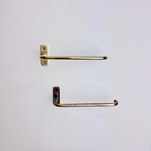 May include: Two brass towel racks mounted on a white wall. The top rack is a straight bar with a rectangular mounting plate and two screws. The bottom rack has a curved bar and a right-angle mounting plate.