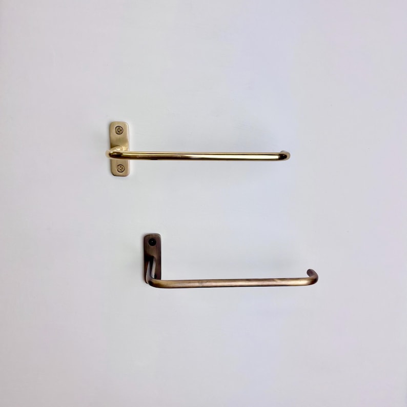 May include: Two brass towel racks mounted on a white wall. The top rack is a straight bar with a rectangular mounting plate and two screws. The bottom rack has a curved bar and a right-angle mounting plate.
