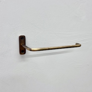 May include: A brass-toned towel bar mounted on a white wall. The bar has a rectangular base with two screw holes and a curved, horizontal arm. The metal has a slightly aged appearance.