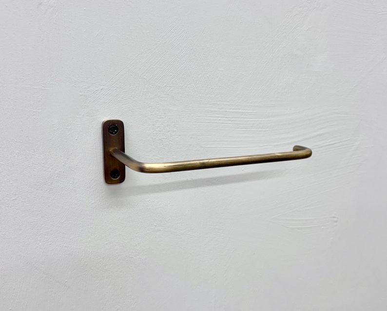 May include: A brass-toned towel bar mounted on a white wall. The bar has a rectangular base with two screw holes and a curved, horizontal arm. The metal has a slightly aged appearance.