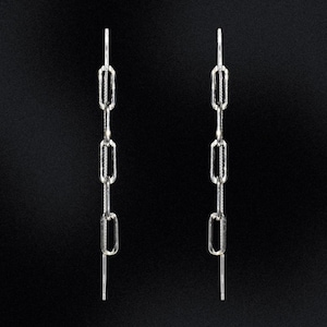 May include: A pair of silver chain earrings. Each earring has a series of elongated, rectangular links connected vertically, with a thin post at the top for insertion and a dangling bar at the bottom. The earrings are set against a black background.