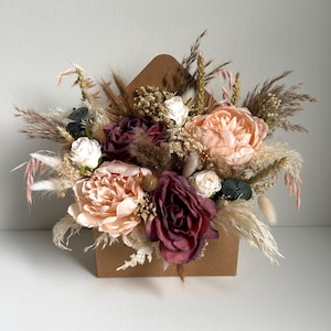 Può includere: Un'arrangiamento floreale con fiori color pesca, bordeaux e bianchi, tra cui rose e peonie, disposti in una busta di carta marrone. Il bouquet è impreziosito da erbe secche e verde, creando un'esposizione rustica ed elegante.