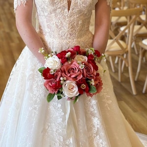 Puede incluir: Una novia con un vestido de novia de encaje blanco sosteniendo un ramo de rosas rojas, rosas y blancas. El ramo está acentuado con vegetación y pequeñas flores blancas. El vestido tiene un escote en V y mangas cortas de encaje.