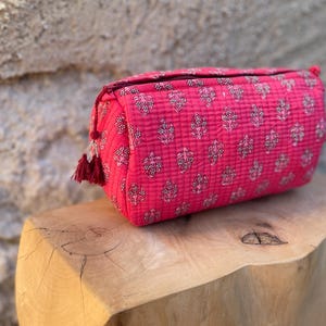Peut inclure: Une trousse de toilette matelassée rectangulaire d'un rose vif, ornée d'un motif floral répétitif imprimé en blanc et noir. La trousse présente une fermeture éclair sur le dessus avec une tirette à pompon rouge foncé et une forme structurée.