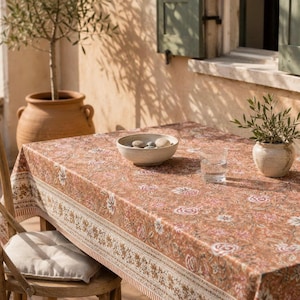 Op de afbeelding: Een rechthoekig tafelkleed met een warme terracotta achtergrond en een herhalend bloemenpatroon in crème, roze en bruine tinten. De stof heeft een decoratieve rand langs de zijkant en ligt over een tafel met een kom en een glas.