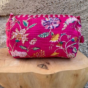 Puede incluir: Un estuche acolchado rectangular de tela rosa fucsia vibrante con un estampado floral multicolor con detalles en amarillo, blanco y verde. El bolso tiene un cierre de cremallera superior y una superficie texturizada y cosida en el exterior.