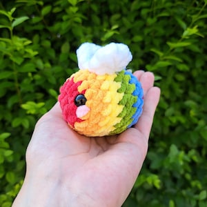 Könnte beinhalten: Ein kleines gehäkeltes Bienentier mit einem regenbogenfarbenen Körper und weißen Flügeln. Die Biene hat ein schwarzes Auge und eine rosa Nase.