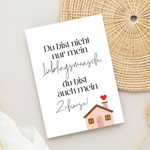 Könnte beinhalten: Eine weiße Karte mit schwarzem Text, der lautet: "Du bist nicht nur mein Lieblingsmensch, du bist auch mein Zuhause!" Darunter ist ein rosa Haus mit einem roten Herzen auf dem Schornstein abgebildet.