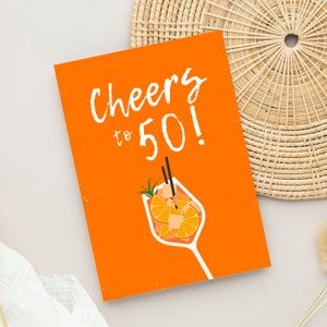 May include: Orange card with the text "Cheers to 50!" in white. A glass of orange juice with a sprig of rosemary and two straws is pictured below the text.