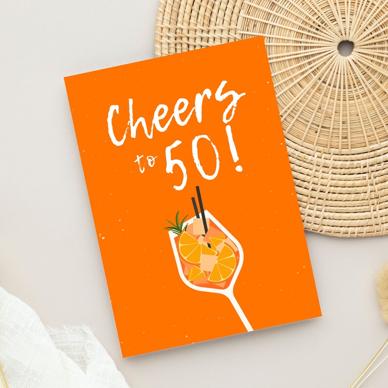 May include: Orange card with the text "Cheers to 50!" in white. A glass of orange juice with a sprig of rosemary and two straws is pictured below the text.