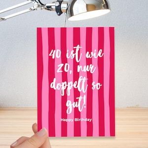 Könnte beinhalten: Eine Geburtstagskarte mit einem rosa-roten Streifenhintergrund. Die Karte trägt den deutschen Text "40 ist wie 20, nur doppelt so gut!" und "Happy Birthday" in weißer Schrift. Die Karte wird von einer Hand gehalten.