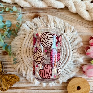 May include: A clear glass with a pink and white pattern of concha pastries. The glass is sitting on a white crocheted coaster.