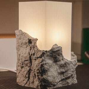 May include: A table lamp featuring a rectangular, vertically ribbed white translucent shade partially encased in a textured, gray, rock-like base. The light emits a warm glow from within the shade, contrasting with the rugged, stone-inspired exterior.