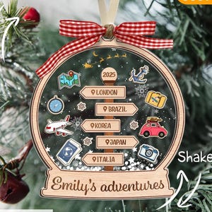 May include: A personalized wooden Christmas ornament shaped like a snow globe, featuring travel-themed icons and destination signs. The ornament has a red and white checkered ribbon and the text "Emily's adventures". The word "CUSTOMIZED" is at the top.