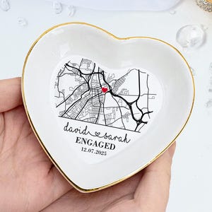 May include: White heart-shaped ceramic ring dish with a gold rim. The dish features a map design with a red heart marking a location. Text on the dish reads "david & sarah ENGAGED 12.07.2025". The word "CUSTOMIZED" is in the top right corner.