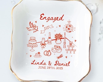 May include: A white ceramic ring dish with a gold rim, featuring a red illustration of engagement-themed icons. The dish is inscribed with the word "Engaged", names, and a date.