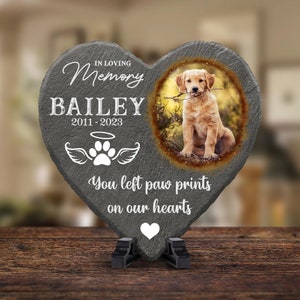 Pu&ograve; includere: Un memoriale in ardesia grigia a forma di cuore con una foto di un cucciolo di golden retriever. L'ardesia ha il testo "In loving memory Bailey 2011-2023" e "You left paw prints on our hearts".