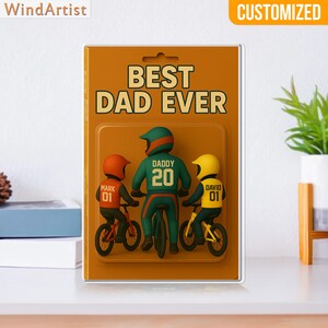 May include: A customized framed art piece with the text "BEST DAD EVER." The artwork depicts a father and two children riding bicycles. The father wears a green and orange cycling outfit, and the children wear orange and yellow outfits. The artwork is displayed on a white surface.