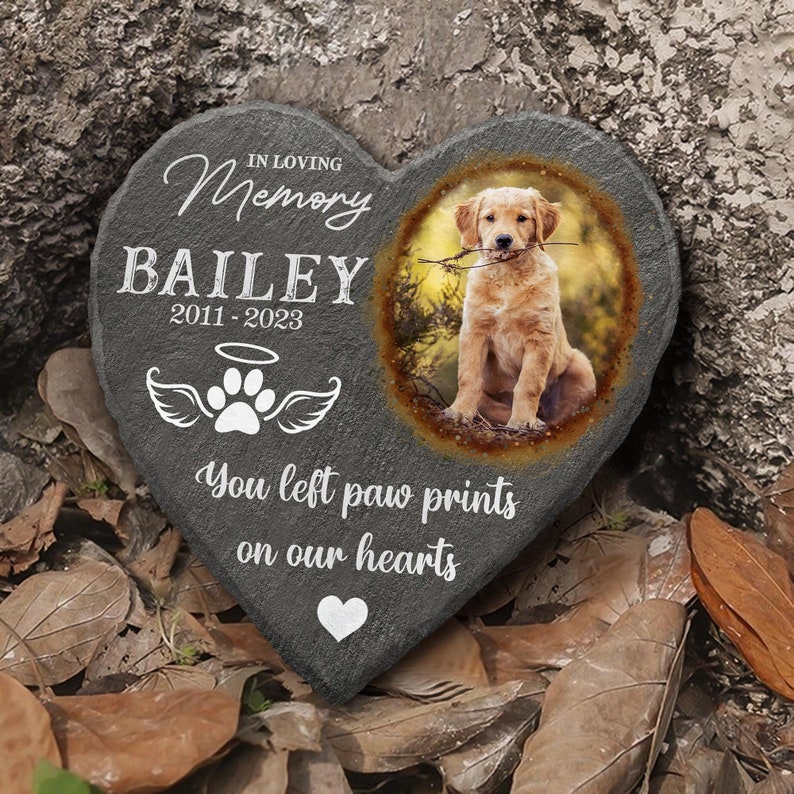 Pu&ograve; includere: Un memoriale in pietra a forma di cuore grigio con una foto di un cucciolo di golden retriever. La pietra ha il testo "In loving memory Bailey 2011-2023" e "You left paw prints on our hearts".