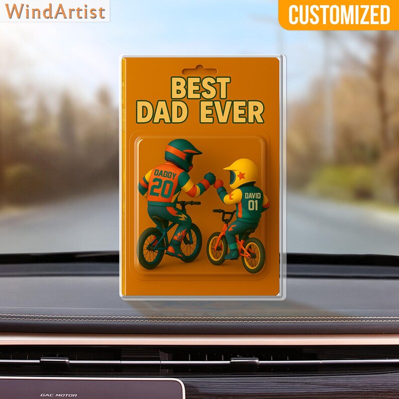 May include: A customized car air freshener with the text "BEST DAD EVER." The design features two cartoon figures on bicycles, one with "DADDY 20" and the other with "DAVID 01." The background is a blurred outdoor scene.