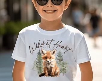 Pode incluir: Camiseta branca com uma ilustração em aquarela de uma raposa e pinheiros. O texto "Wild at Heart" está escrito acima da raposa em itálico. A camiseta está sendo usada por uma criança.