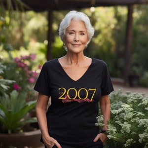 Peut inclure: Une femme portant un t-shirt noir &agrave; col en V avec le texte "EST 2007" en lettres dor&eacute;es et un motif rouge et or.